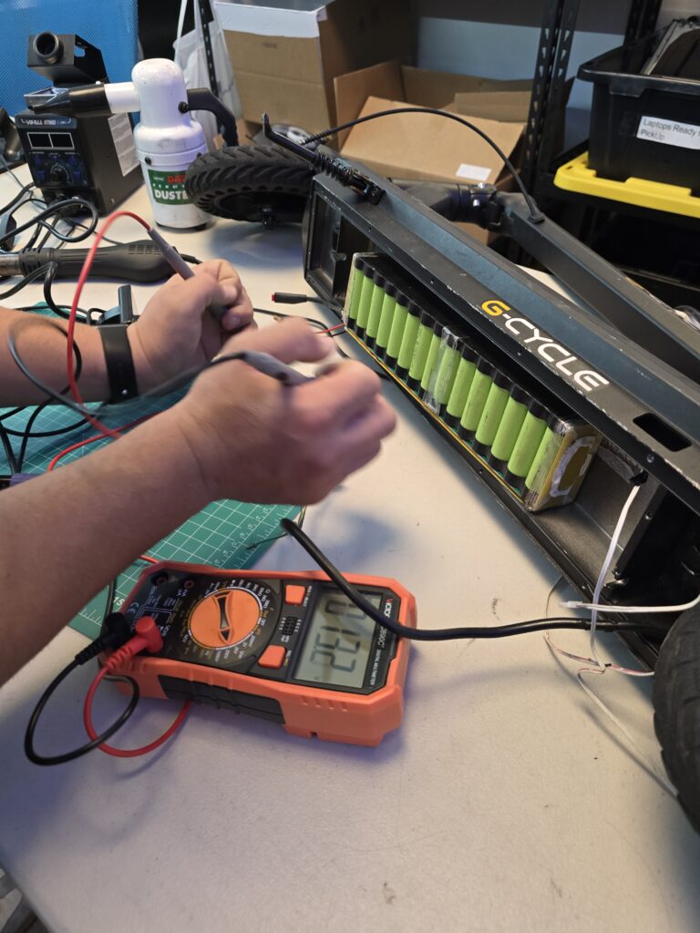 Technician testing a G-Cycle electric scooter battery with a multimeter at Techy Doral